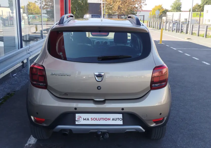 Vue arrière d'une Dacia Sandero Stepway beige dune de 2020 avec attelage et feux arrière foncés sur parking.