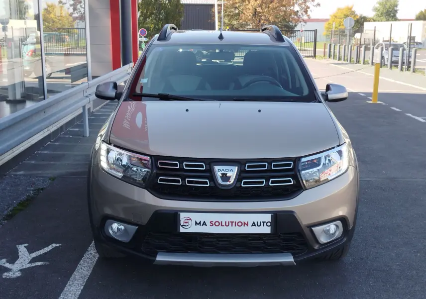Vue frontale d'une Dacia Sandero Stepway beige dune 2020 avec calandre noire et phares allumés.