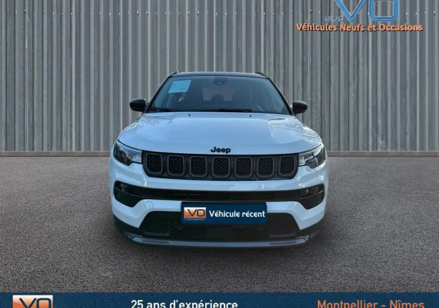 Vue frontale d'un Jeep Compass blanc 2023 avec calandre noire et phares LED, stationné devant un mur métallique.