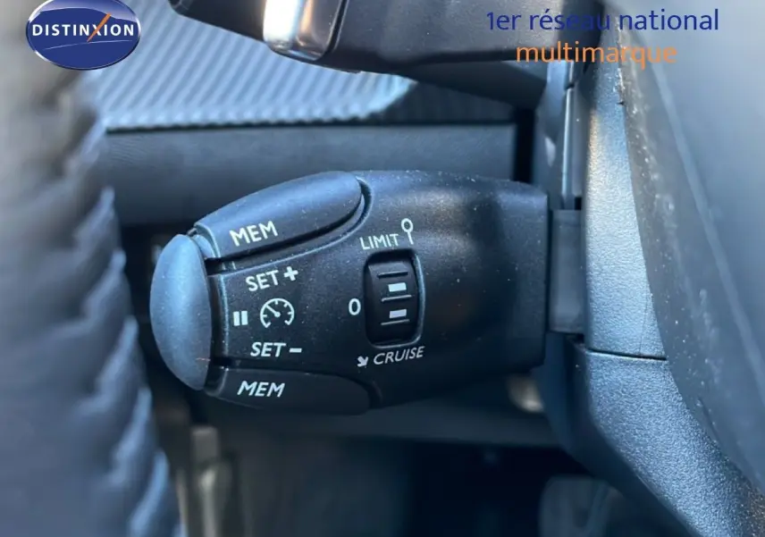 Gros plan sur la commande de régulateur de vitesse noire à gauche du volant dans une Peugeot 208 noire hybride 2025.