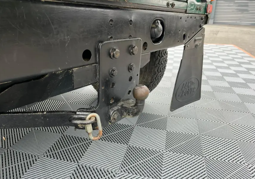 Gros plan sur l'attelage arrière noir et le bavette Land Rover d'un Defender 90 vert, vue côté gauche.