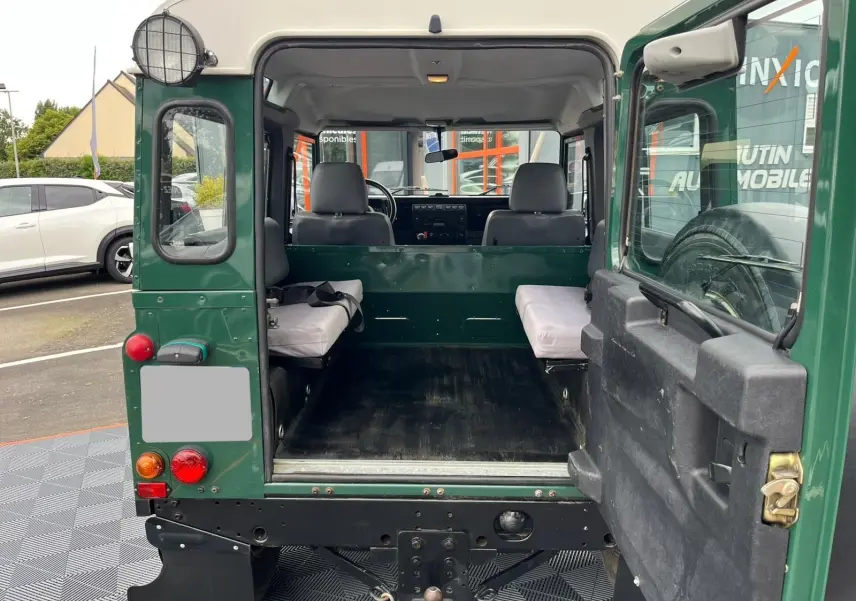 Vue arrière ouverte du Land Rover Defender 90 vert 2004 montrant l'intérieur utilitaire avec banquettes latérales et roue de secours.