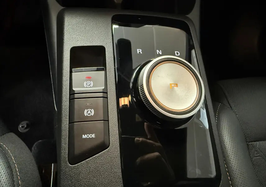 Gros plan sur la molette de boîte automatique et les boutons de contrôle du frein à main dans l’habitacle noir du MG3 Hybrid+ Luxury.