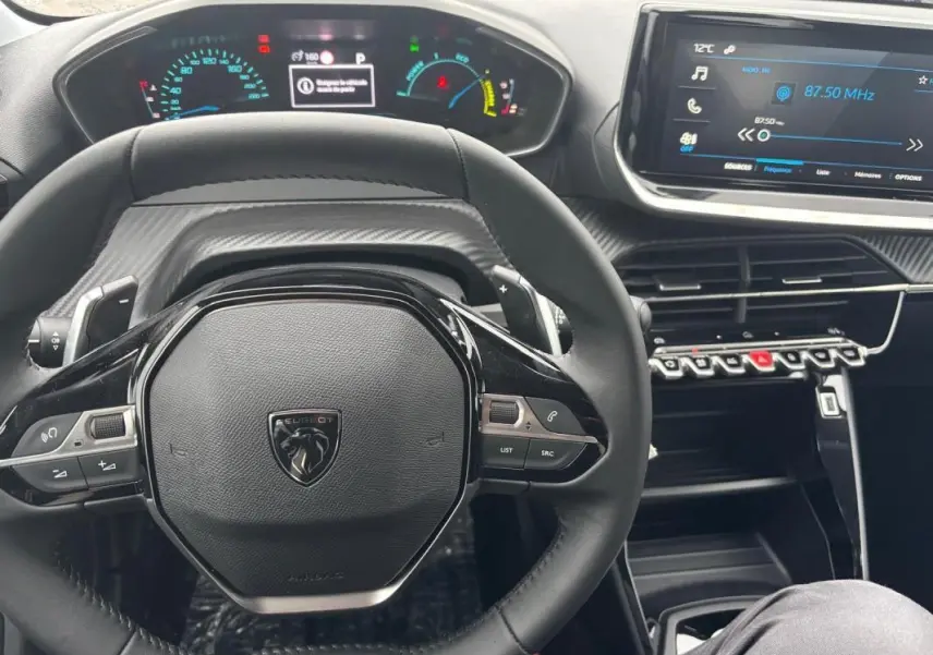 Vue intérieure centrée sur le volant noir de la Peugeot 208 Hybrid 100 e-DCS6 Style avec tableau de bord numérique et écran tactile.