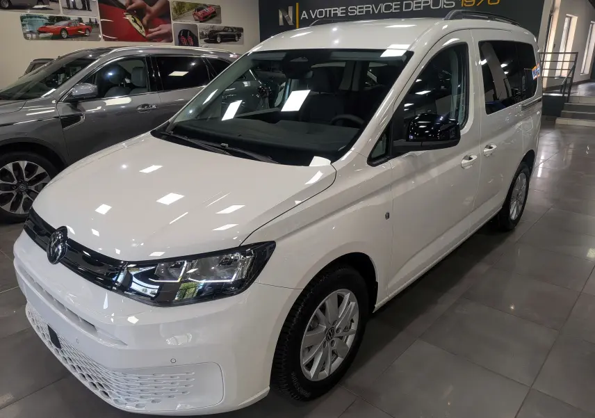 Vue 3/4 avant droite d’un Volkswagen Caddy blanc 2025 en showroom avec vitres teintées et jantes alliage.