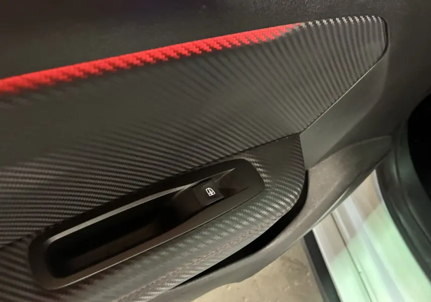 Gros plan sur la commande de lève-vitre côté conducteur avec finition carbone et surpiqûres rouges dans une Renault Mégane IV RS 280 blanche.