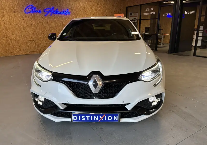 Renault Mégane IV RS 280 blanc vue de face, phares allumés et calandre noire distinctive en intérieur showroom.