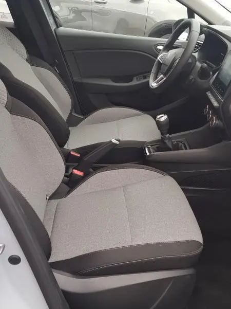 Vue intérieure côté conducteur de la Renault Clio gris 2025, avec sièges tissu clair et levier de vitesses manuel visible.