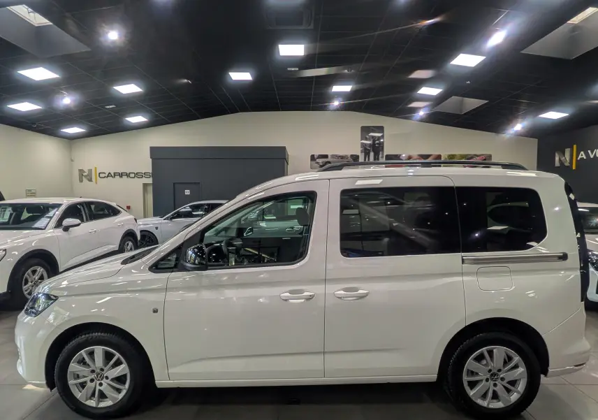 Vue de profil côté gauche d'un Volkswagen Caddy blanc 2025 en intérieur, avec vitres arrière teintées et barres de toit.