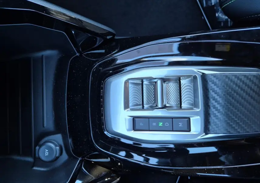 Gros plan sur la console centrale noire brillante de la Peugeot 208 Hybrid 136 e-DCS6 GT avec commandes de boîte auto et prise 12V.