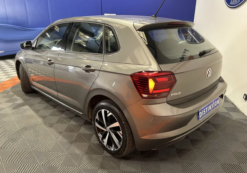 Volkswagen Polo gris vue 3/4 arrière droit, feux arrière allumés et jantes noires à motifs argentés.