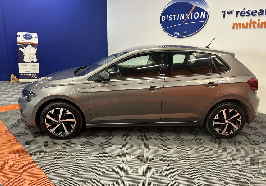 Vue de profil côté gauche d'une Volkswagen Polo gris métallisé 2018, avec jantes alliage et vitres teintées en arrière-plan showroom.