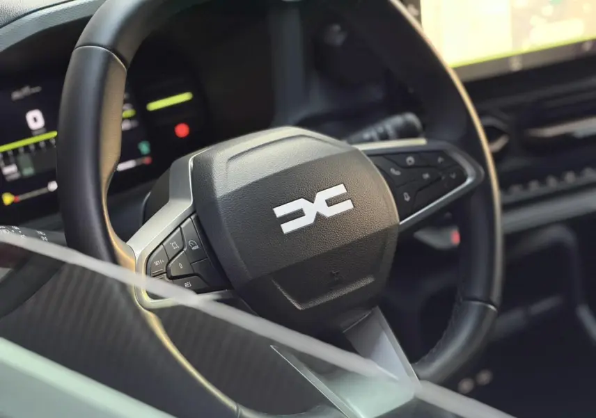 Gros plan sur le volant noir avec logo Dacia stylisé, tableau de bord digital en arrière-plan flou.