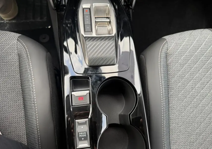 Vue plongeante sur la console centrale noire brillante de la Peugeot 208 Hybrid 145, avec levier de vitesse et porte-gobelets.