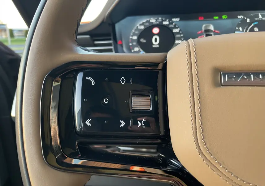 Gros plan sur les commandes gauche du volant beige du Range Rover Sport P440 HSE R-Dynamic 2023, affichage numérique en arrière-plan.