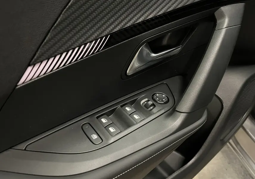 Gros plan sur la commande des vitres et la poignée intérieure côté conducteur du Peugeot 2008 gris, avec finition carbone et surpiqûres blanches.