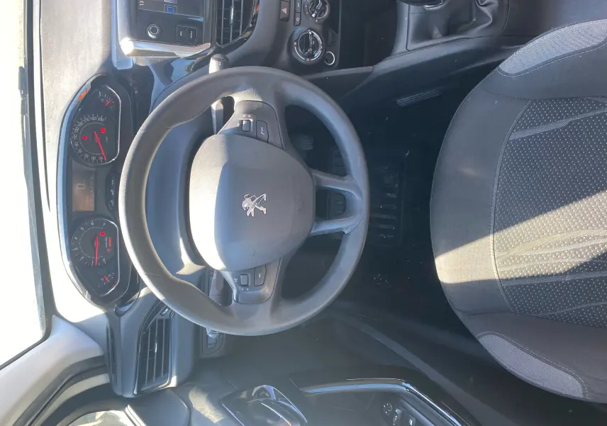 Vue intérieure centrée sur le volant et tableau de bord d'une Peugeot 208 blanche, avec sièges noirs et commandes visibles.