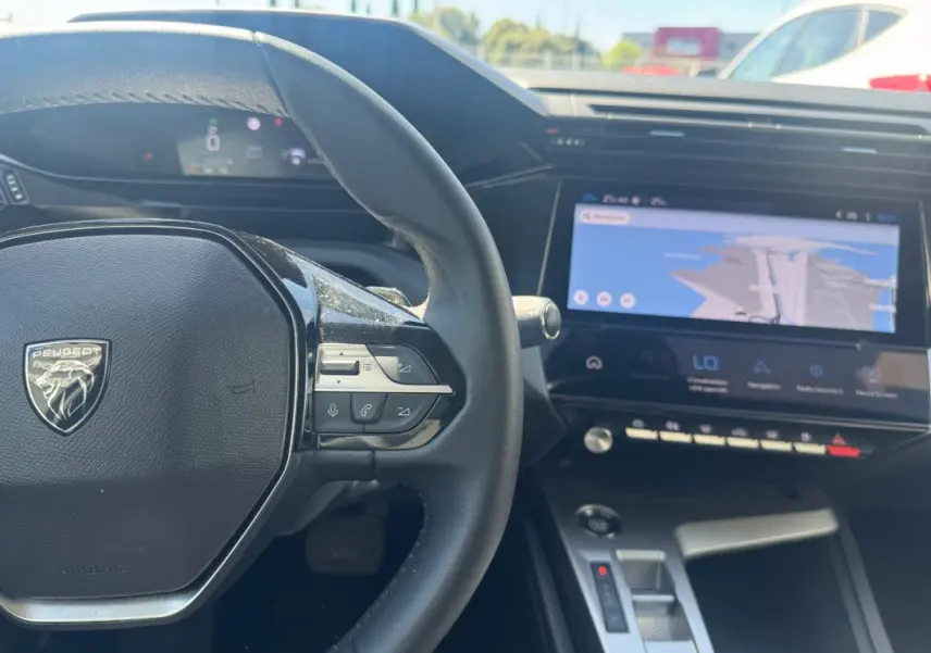 Vue intérieure centrée sur le volant Peugeot gris et le grand écran tactile du tableau de bord avec navigation affichée.