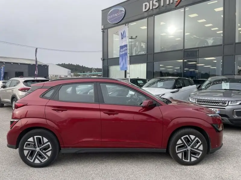Profil côté gauche d'un Hyundai Kona électrique rouge métal 2021 garé devant un showroom avec jantes distinctives.