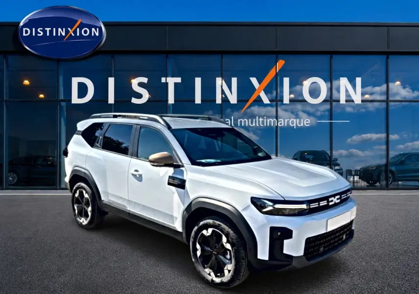 Dacia Bigster blanc en 3/4 avant droit, avec barres de toit et jantes alu distinctives, devant vitrine Distinxion.