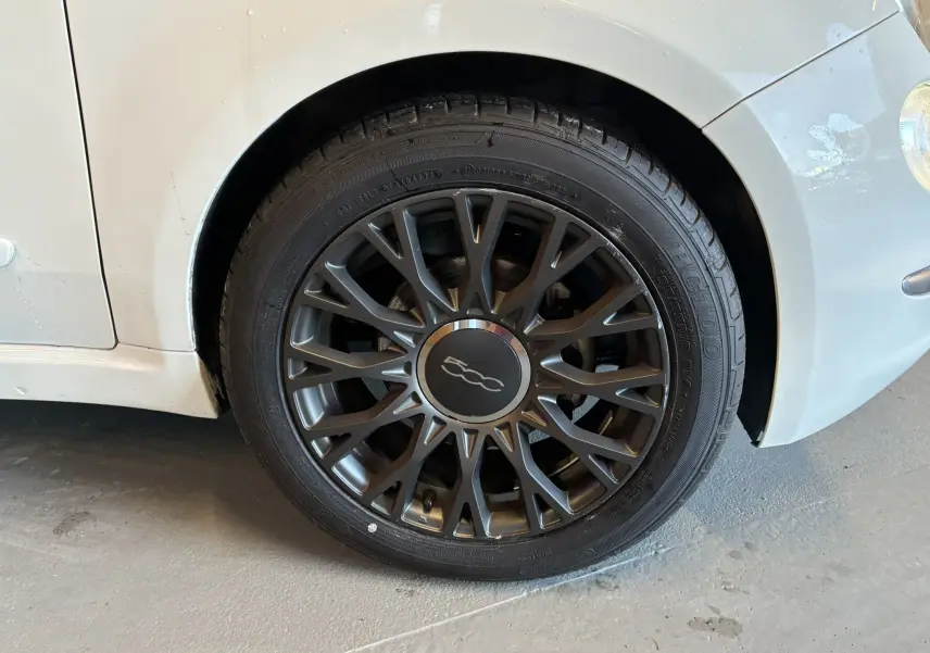 Gros plan sur la roue avant gauche d'une FIAT 500 blanche, mettant en valeur la jante noire au design complexe.