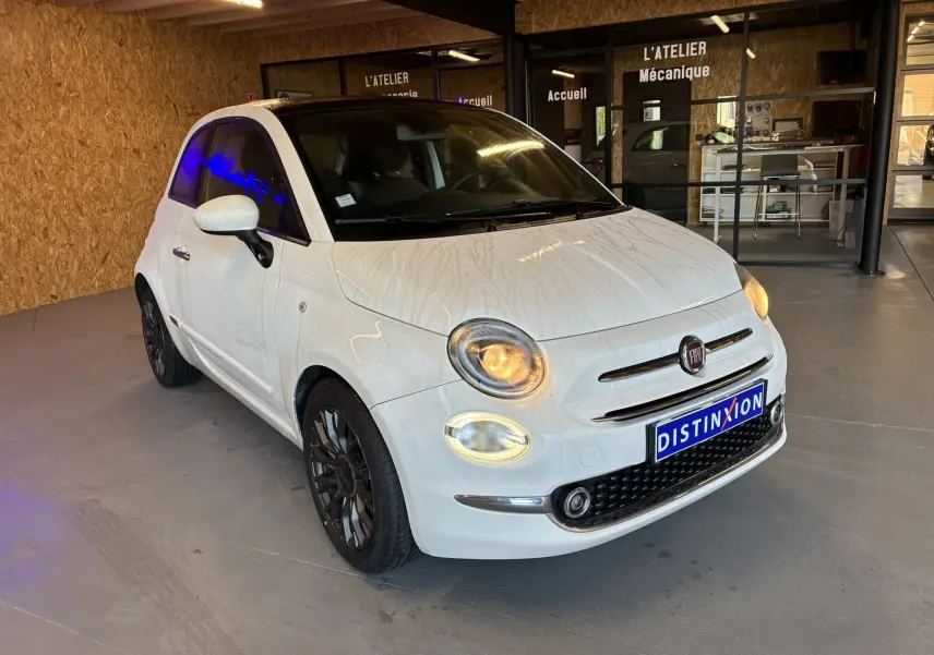 Vue 3/4 avant droite d'une Fiat 500 blanche de 2021 avec phares allumés, stationnée en intérieur devant un atelier mécanique.