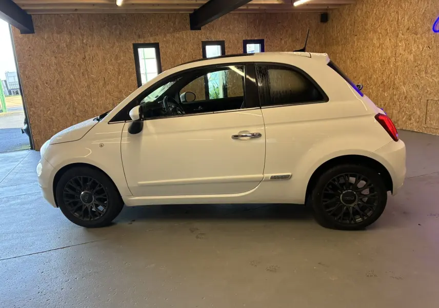 Profil droit d'une FIAT 500 blanche de 2021 avec jantes noires et intérieur visible en arrière-plan bois.