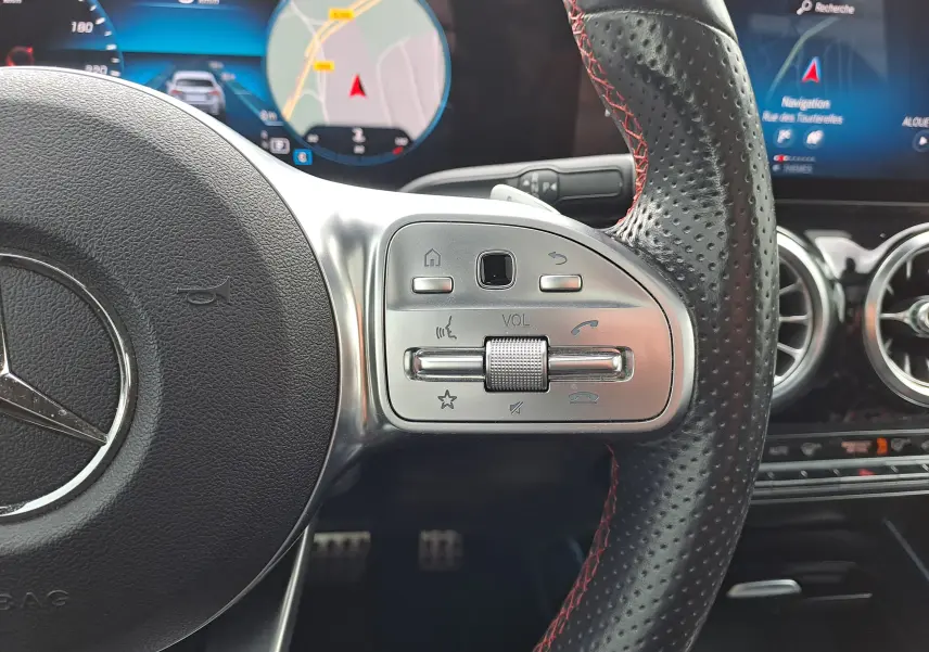 Gros plan sur le volant perforé noir avec surpiqûres rouges d'une Mercedes Classe B AMG Line 2019, affichage numérique en arrière-plan.