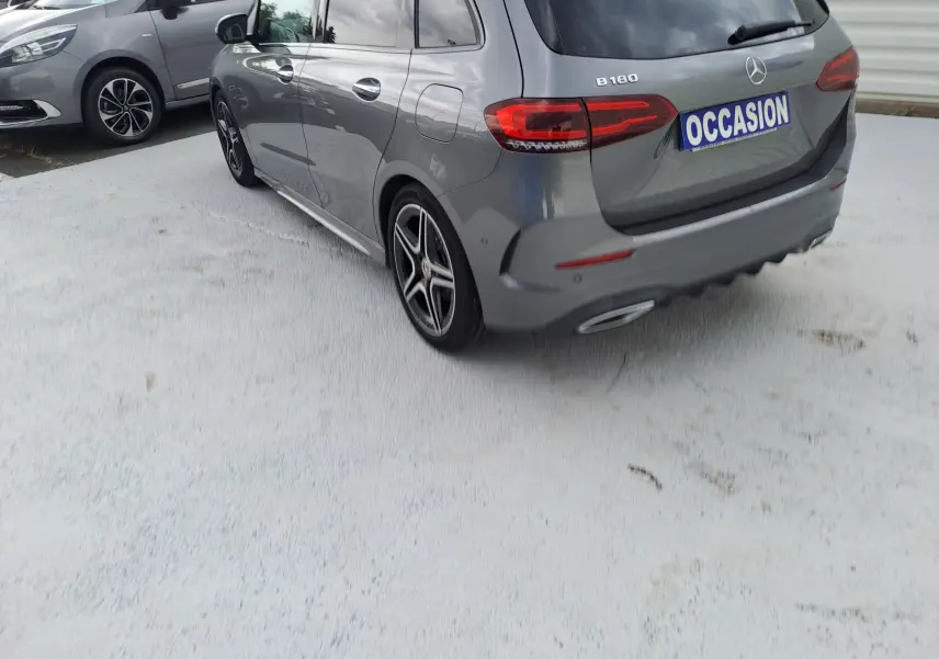 Vue 3/4 arrière droite d'une Mercedes Classe B gris foncé métallisé avec jantes AMG et badge B180 visible.