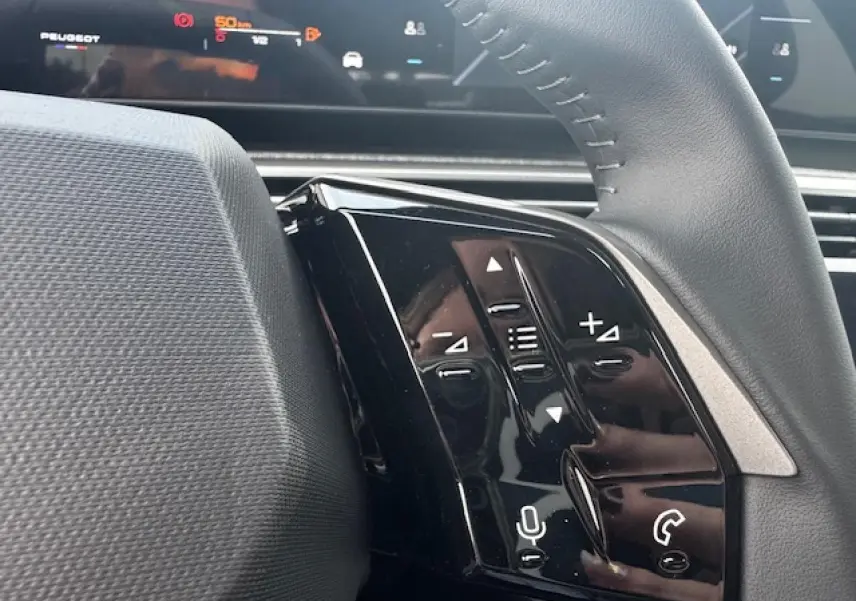 Gros plan sur les commandes gauche du volant cuir noir du Peugeot 5008 bleu Ingaro, avec écran numérique en arrière-plan.