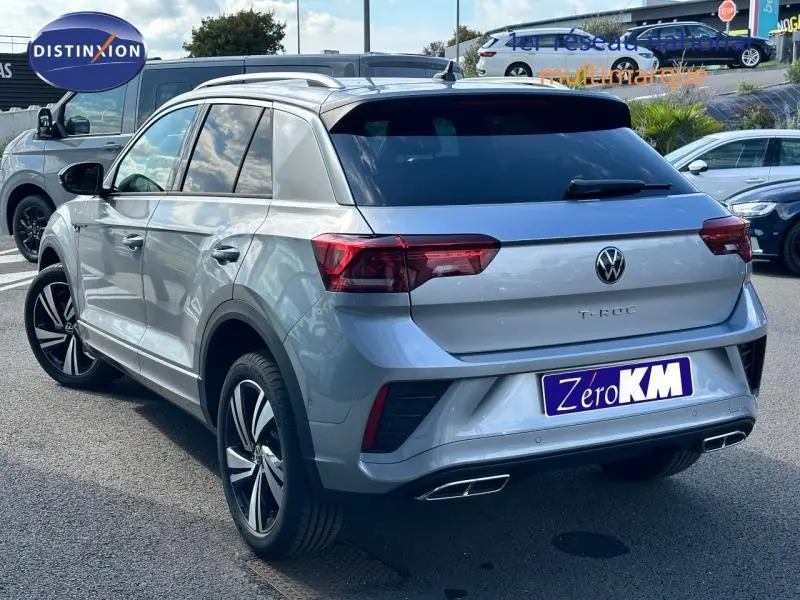 Volkswagen T-Roc 2025 gris clair en vue 3/4 arrière droit, avec jantes alu et double sortie d'échappement chromée.