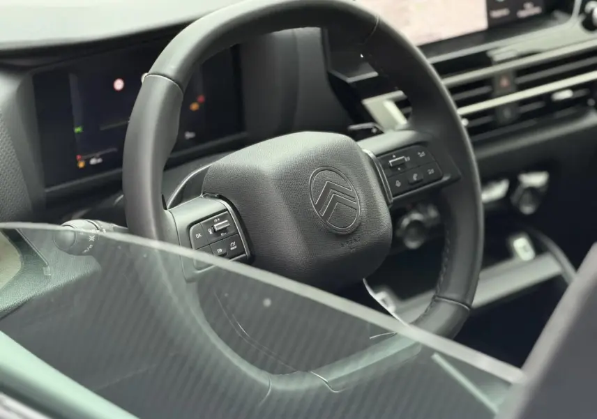 Vue rapprochée du volant noir de la Citroën C4 2025, avec commandes intégrées et tableau de bord numérique flou en arrière-plan.