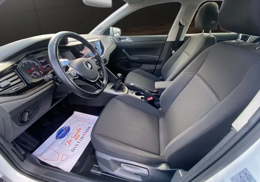 Vue intérieure côté conducteur de la Volkswagen Polo blanche, montrant les sièges noirs et le tableau de bord avec boîte manuelle.