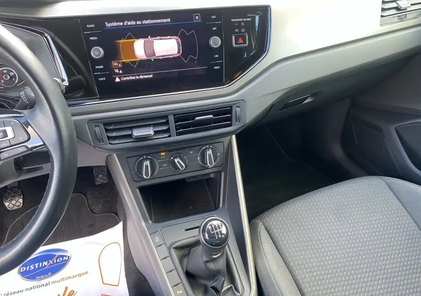 Vue intérieure côté conducteur de la Volkswagen Polo blanche, avec tableau de bord et levier de vitesses manuel en évidence.