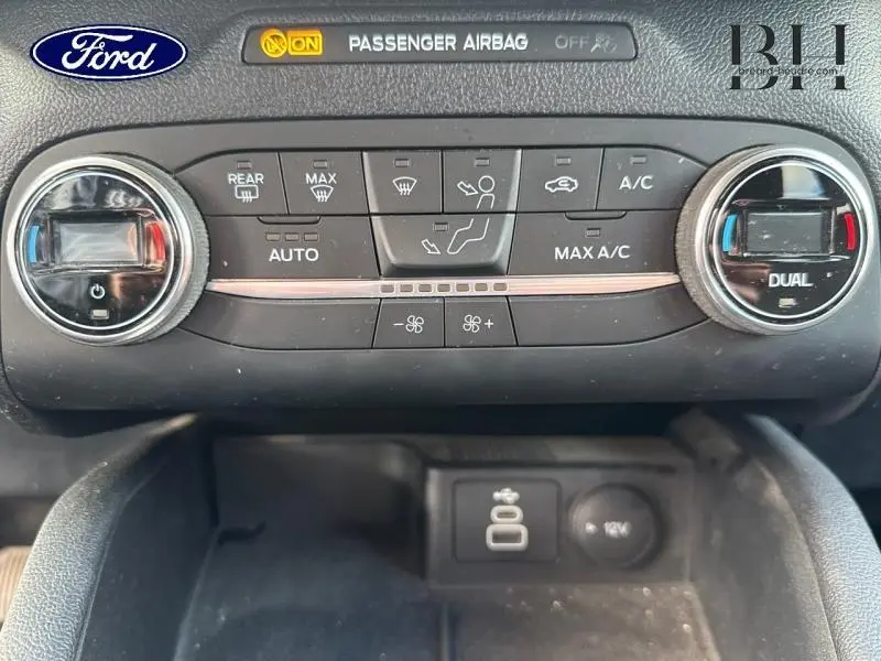 Vue rapprochée du tableau de bord noir du Ford Kuga 2022, montrant les commandes de climatisation et l'indicateur airbag passager.