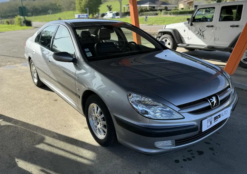 Peugeot 607 gris vue 3/4 avant droit, capot brillant et jantes alliage sous un abri en extérieur.