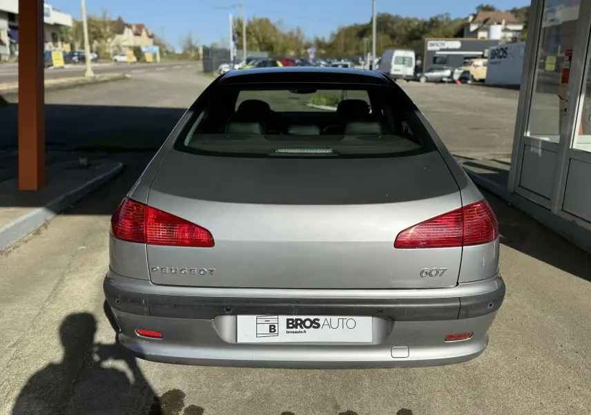 Vue arrière d'une Peugeot 607 gris métallisé garée devant un bâtiment commercial par temps ensoleillé.