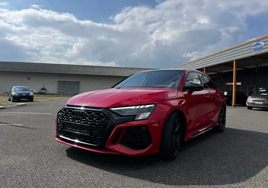 Audi RS3 Sportback rouge vue 3/4 avant droit, calandre noire et jantes noires, stationnée en extérieur.