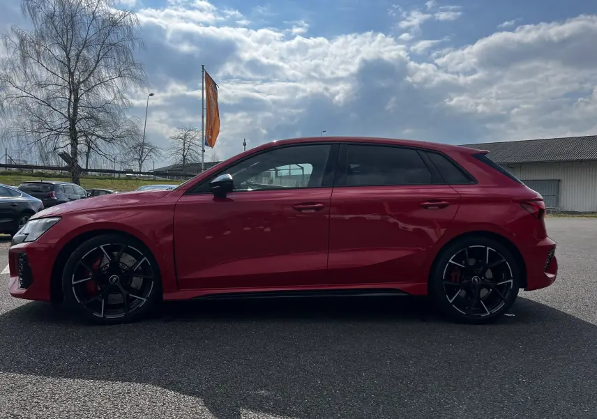 Profil côté gauche d'une Audi RS3 Sportback rouge 2022 avec jantes noires et vitres teintées sur parking extérieur.