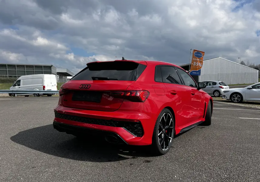 Audi RS3 Sportback rouge vue 3/4 arrière droit sur parking, avec jantes noires et vitres teintées.