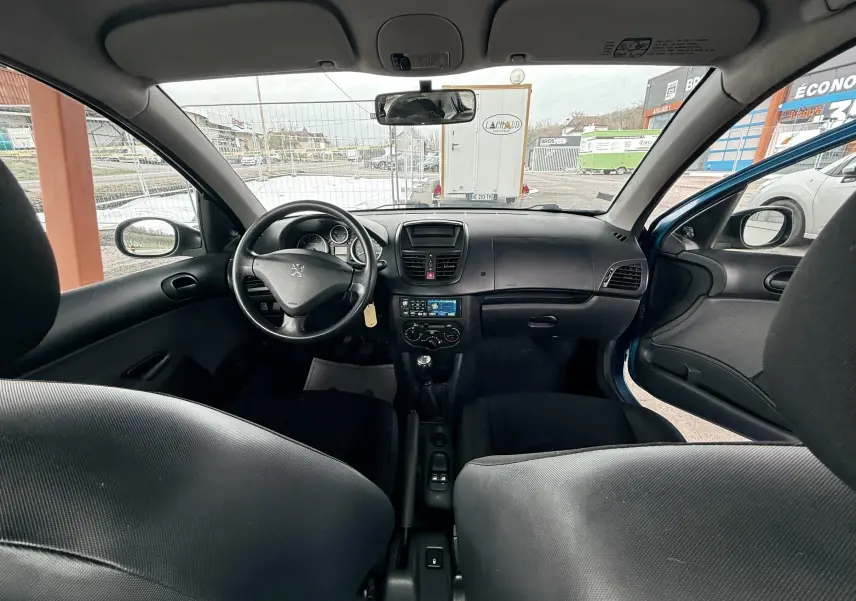 Vue intérieure avant de la Peugeot 206+ bleu, montrant le tableau de bord et le volant côté gauche avec porte ouverte.