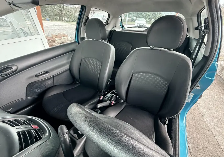 Vue intérieure côté conducteur d’une Peugeot 206+ bleue, montrant les sièges avant en tissu noir et le volant.