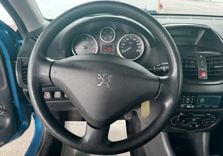 Vue intérieure centrée sur le volant noir de la Peugeot 206+ bleu, avec tableau de bord et commandes visibles.
