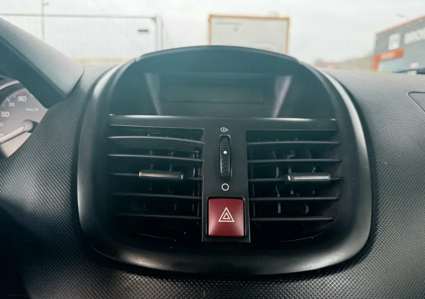 Gros plan sur la sortie d’air centrale noire et le bouton de warning rouge du tableau de bord d’une Peugeot 206+ bleue.