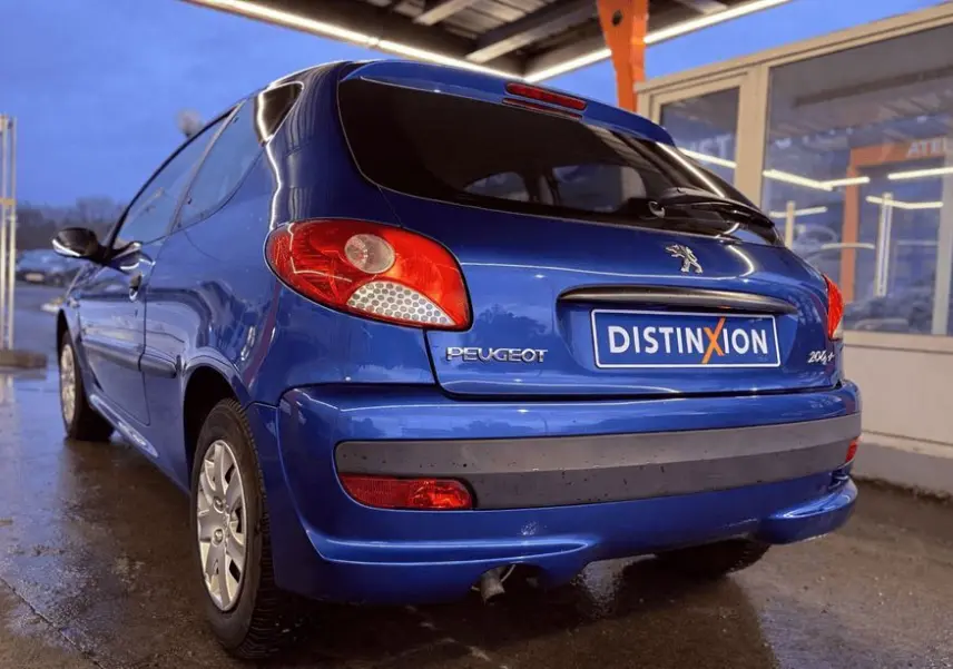 Vue 3/4 arrière droite d'une Peugeot 206+ bleue garée sous un abri avec plaque Distinxion visible.