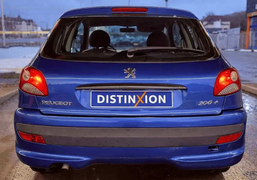 Vue arrière d'une Peugeot 206+ bleue avec plaque Distinxion, feux arrière allumés et hayon fermé.