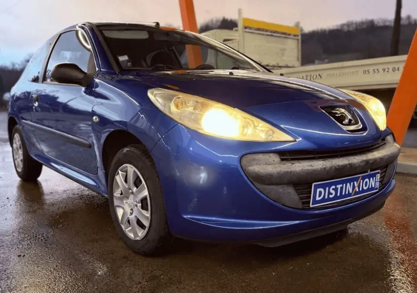 Peugeot 206+ bleu vue 3/4 avant droit avec phares allumés sur sol mouillé, calandre noire et logo visible.