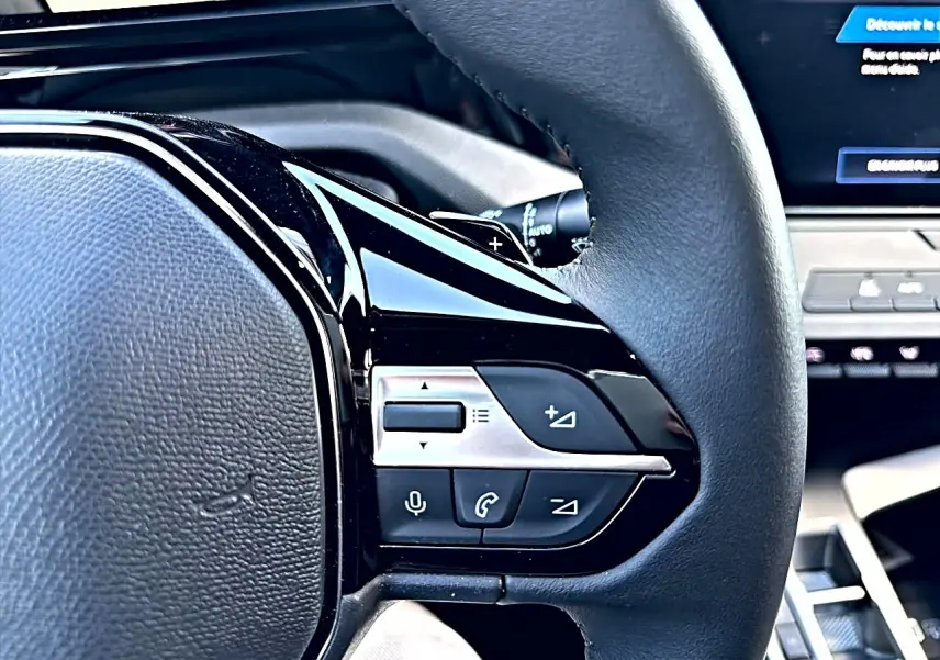 Gros plan sur les commandes multifonctions côté gauche du volant noir du Peugeot 308 gris selenium, avec écran tactile en arrière-plan.