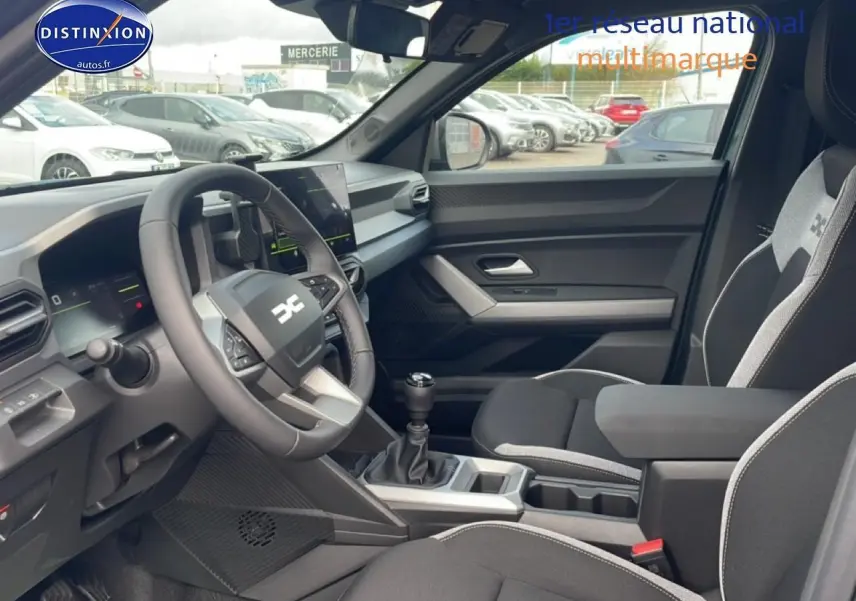 Vue intérieure côté conducteur du Dacia Duster 2026, montrant le volant multifonction, la boîte manuelle et les sièges noirs avec surpiqûres grises.