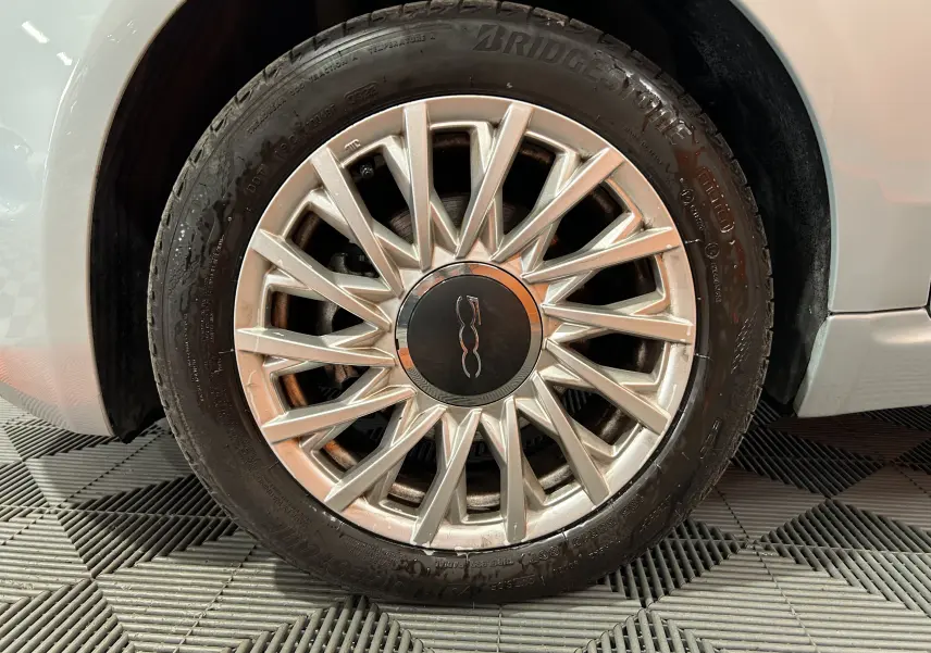 Gros plan sur la roue avant droite d'une FIAT 500 blanche avec jante argentée à motifs rayonnants.